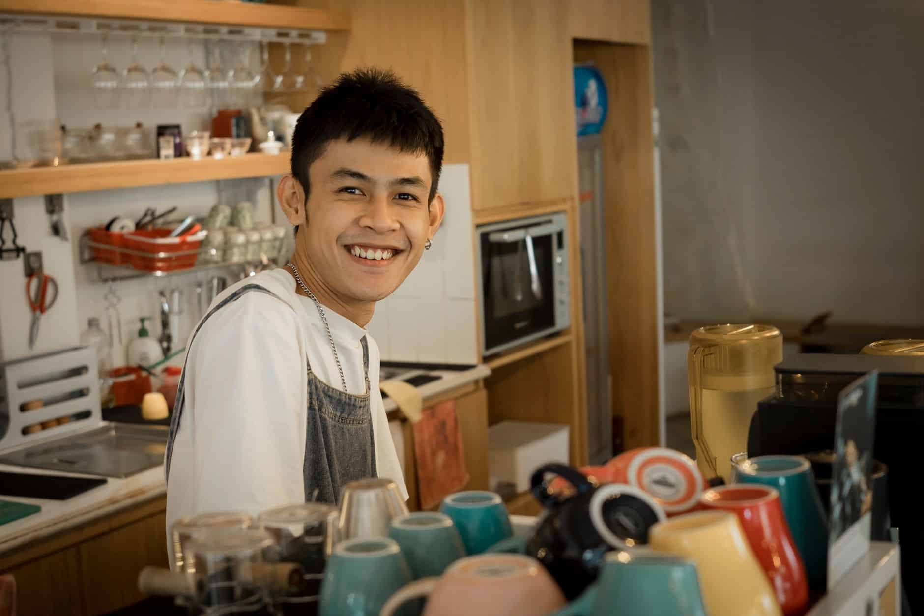 Home Coffee shop marketing: cheerful barista smiling behind counter in a cosy coffee shop with colourful cups